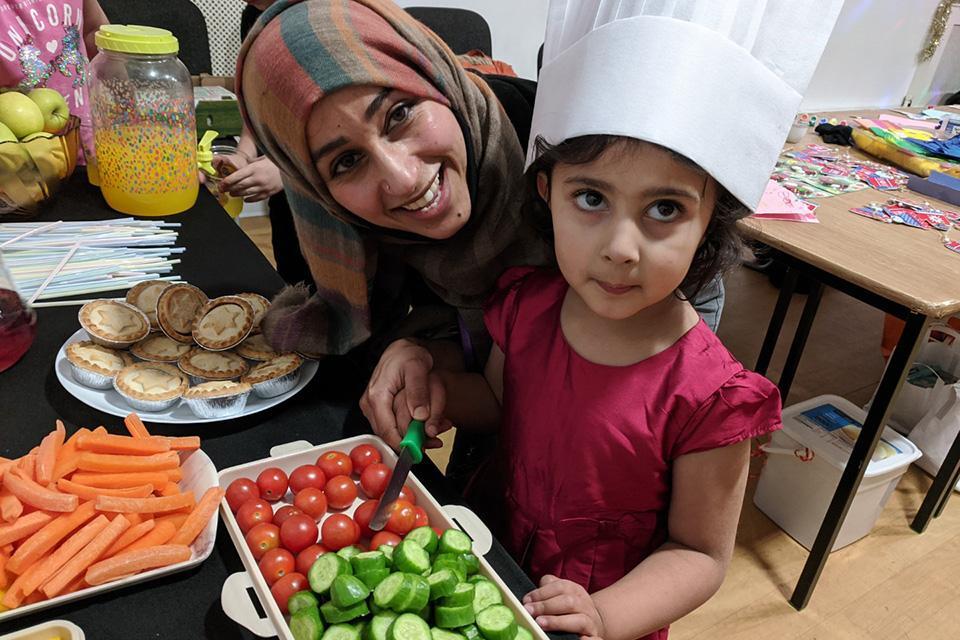 A woman and a child prepare vegetables on a table. They smile to the camera and the child is wearing a white chef's hat.