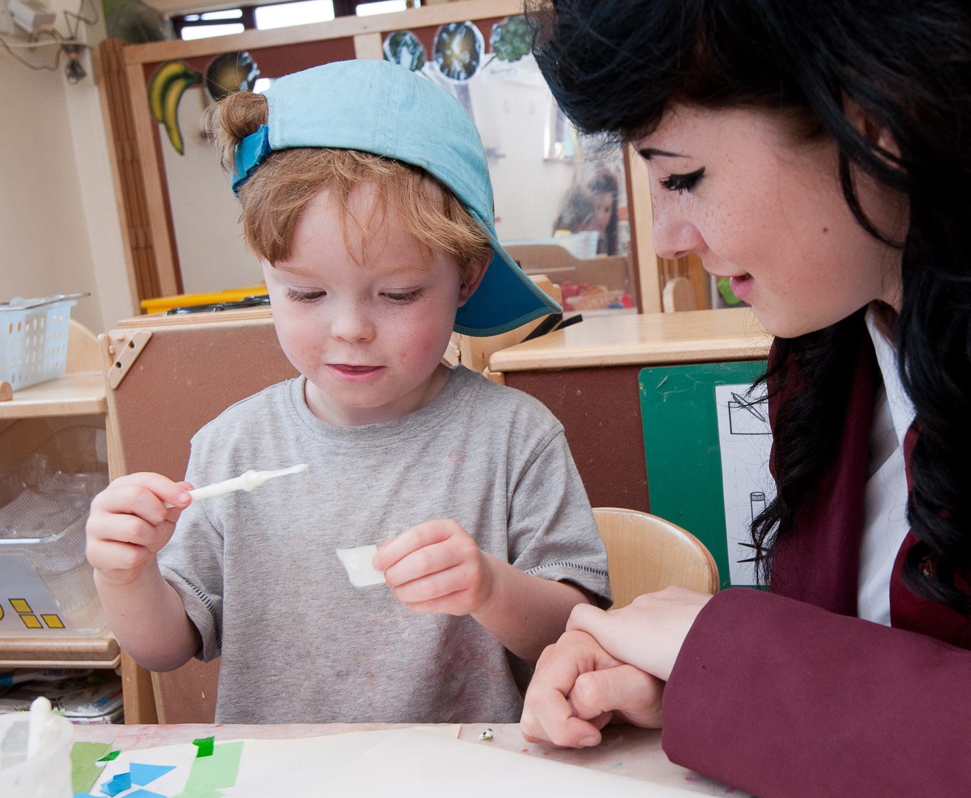 Young boy gluing a piece of paper with a teacher watching him