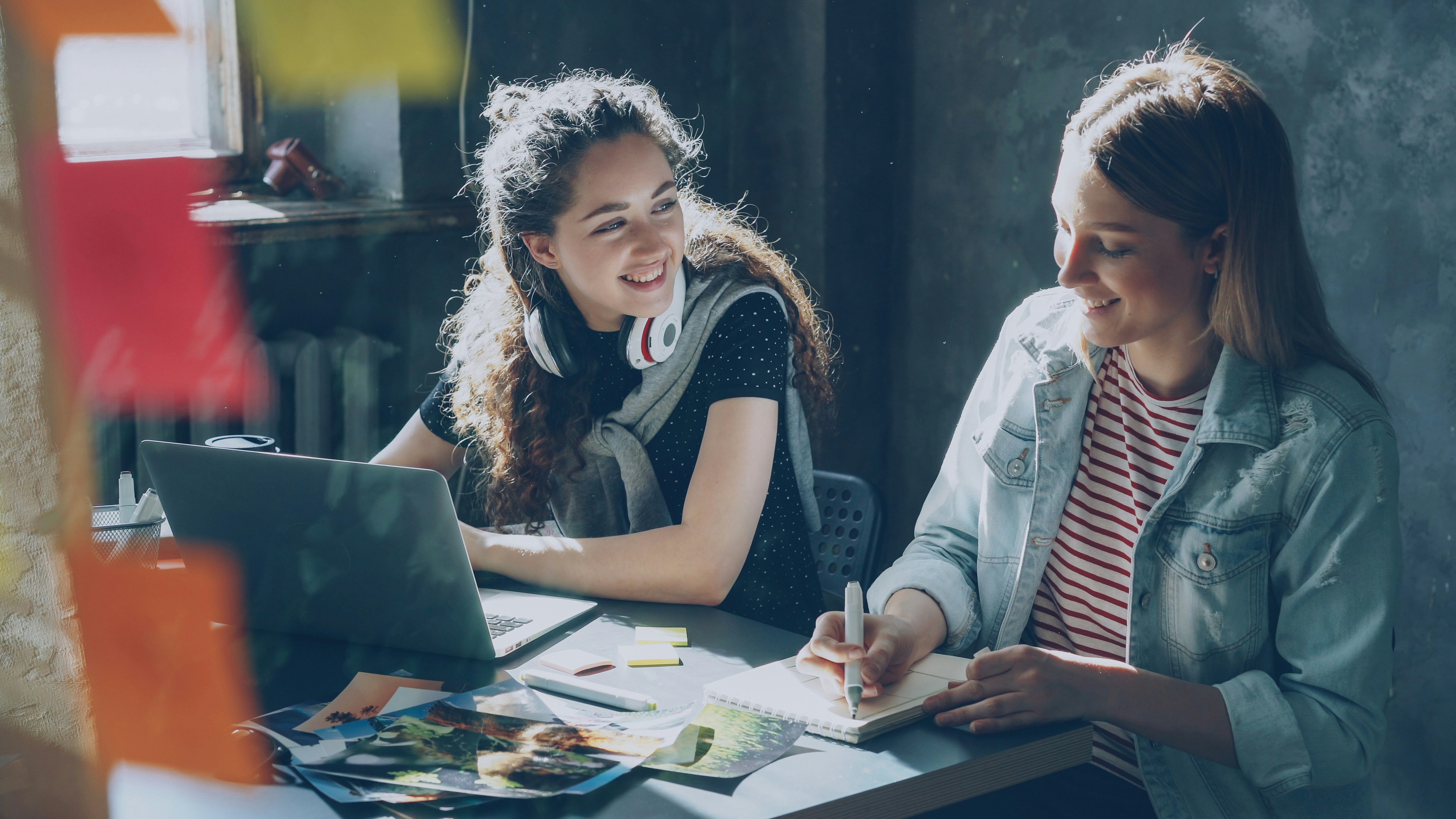 Two women work at a desk, one writes in a notebook and another smiles at her. She has an open laptop in front of her and there are pieces of paper and pens scattered across the desk.